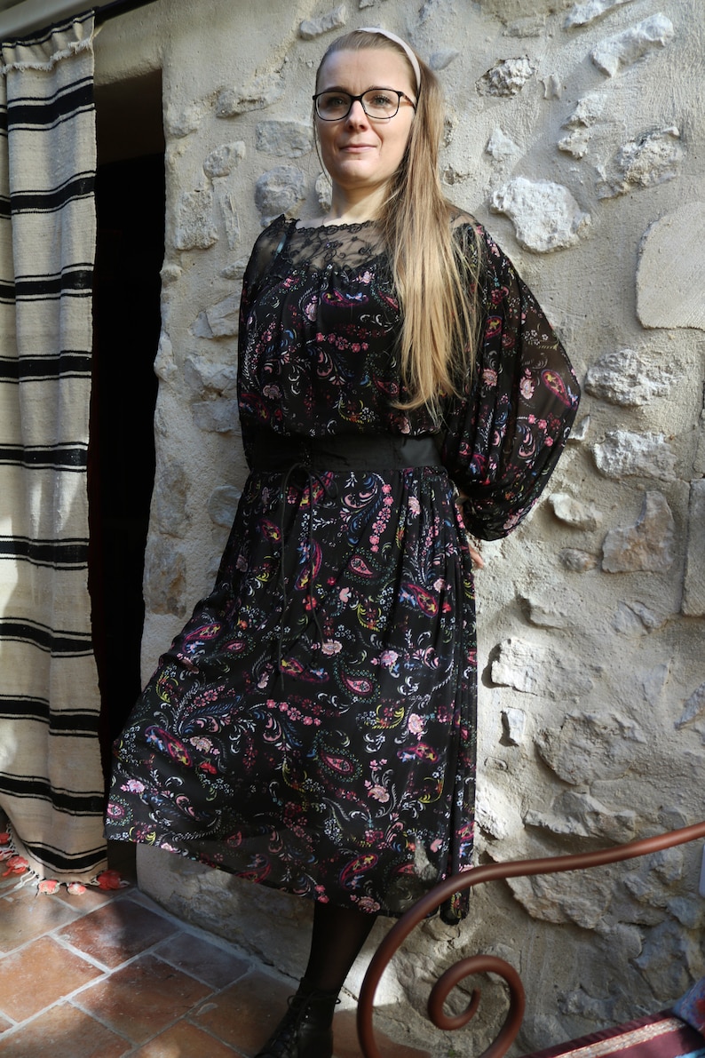 Puede incluir: Una mujer con un vestido negro floral con cintur&oacute;n negro y mangas largas. Est&aacute; de pie frente a una pared de piedra y tiene el pelo largo y rubio. Lleva gafas y una diadema.