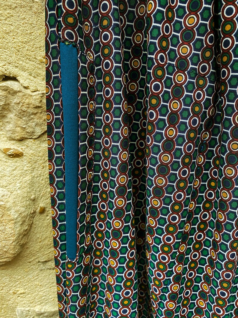 May include: Close-up of a teal blue fabric pocket sewn onto a green, red, white, and yellow patterned fabric. The pattern is made up of circles and hexagons.