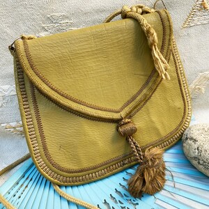 May include: A mustard yellow leather shoulder bag with a braided rope strap and tassel. The bag has a flap closure and is decorated with brown stitching.