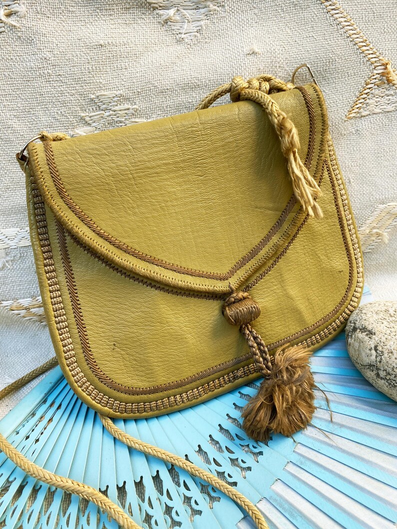 May include: A mustard yellow leather shoulder bag with a braided rope strap and tassel. The bag has a flap closure and is decorated with brown stitching.