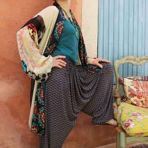 May include: A woman wearing a colorful patterned kimono over a teal top and black and white patterned pants. She is standing in front of a blue door and a chair with a floral cushion.