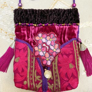 May include: A pink velvet bag with gold embroidery and a floral design. The bag has a purple drawstring and tassels.