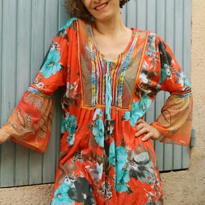 May include: A woman wearing a red and orange floral print top with a turquoise and brown design. The top has a gathered neckline with a turquoise tassel. The woman is wearing green trousers.
