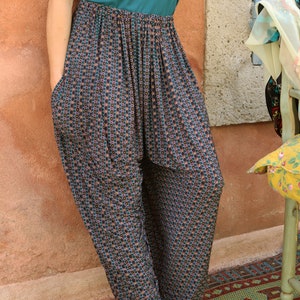 May include: A woman wearing a teal tank top and patterned pants with a green pendant necklace. She is standing in front of a wall and wearing gold sandals.