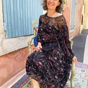 Puede incluir: Una mujer lleva un vestido negro con estampado floral y una superposici&oacute;n de encaje transparente. Est&aacute; sentada en una silla con un coj&iacute;n de estampado floral. La silla est&aacute; delante de una puerta de madera azul y una pared rosa.