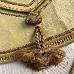 May include: A close-up of a brown leather purse with a brown tassel. The purse has a brown braided strap and a brown leather flap.