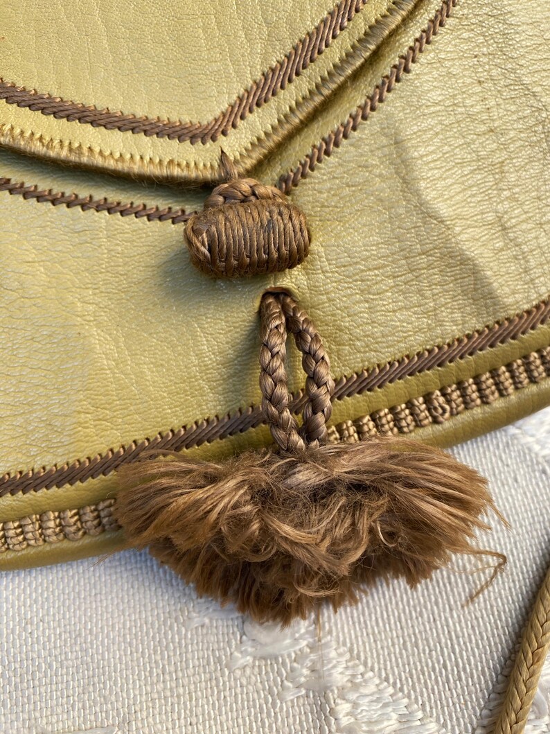 May include: A close-up of a brown leather purse with a brown tassel. The purse has a brown braided strap and a brown leather flap.