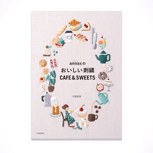 Op de afbeelding: Een boekomslag met een lichtbruine achtergrond en een cirkel van kleurrijke geborduurde afbeeldingen van eten en drinken. De tekst "annasの おいしい刺繍 CAFE & SWEETS" staat in het midden van de cirkel. De tekst "川畑杏奈" staat onder de cirkel.