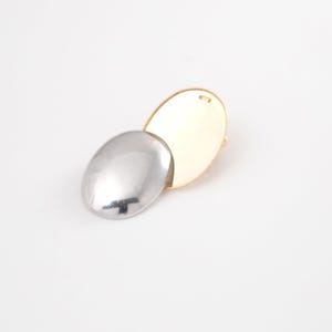May include: A two-tone brooch with an oval silver element overlapping a gold oval. The brooch has a simple, modern design. The background is plain white.