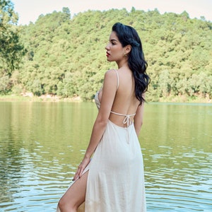 Puede incluir: Una mujer con un vestido maxi blanco con la espalda descubierta y una abertura en el muslo hasta la altura del muslo se encuentra en un lago. El vestido tiene una tira fina y un lazo en la espalda. La mujer mira hacia la izquierda del encuadre.