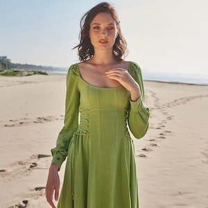 May include: A woman wearing a green, long-sleeved dress with a lace-up bodice. The dress has a square neckline and a flowing skirt. She is standing on a sandy beach with footprints in the sand.