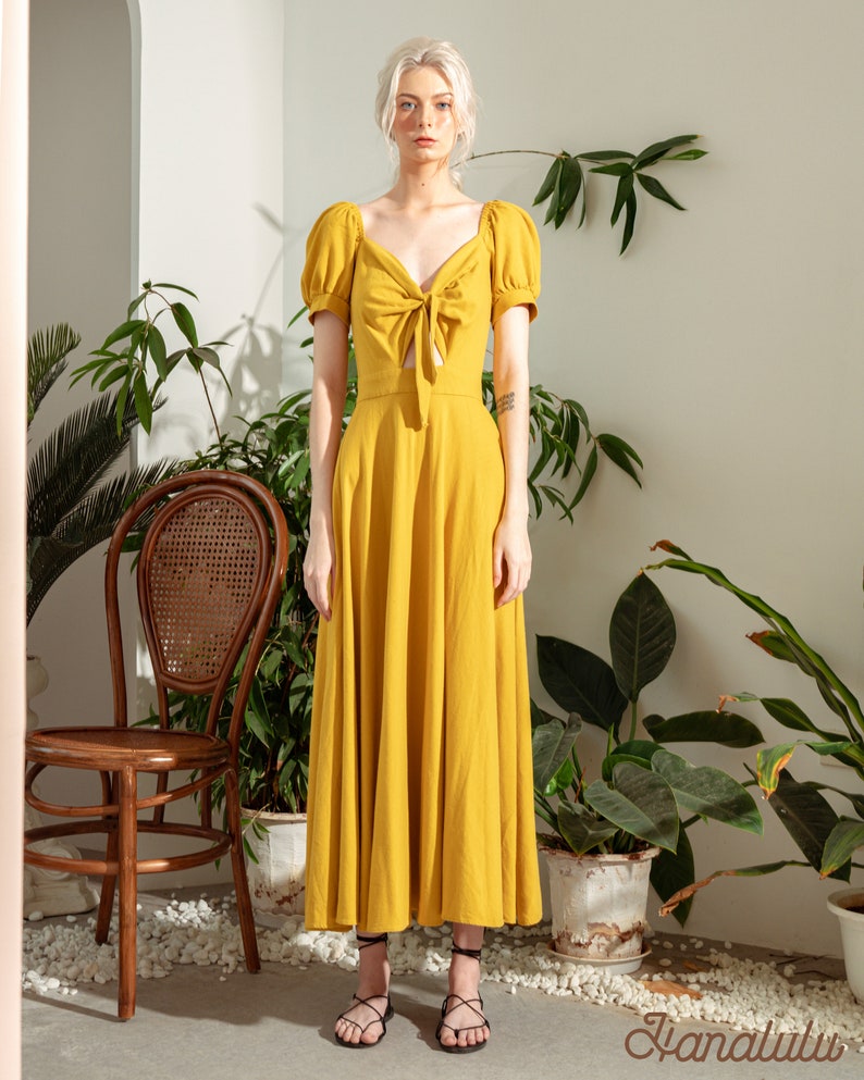 May include: A yellow maxi dress with a tie at the neckline and puff sleeves. The dress is made of a lightweight fabric and has a flowy silhouette.