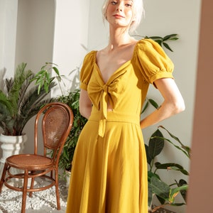 May include: A woman wearing a yellow midi dress with a tie at the neckline and puff sleeves. The dress is made of a lightweight fabric and has a flowy silhouette.