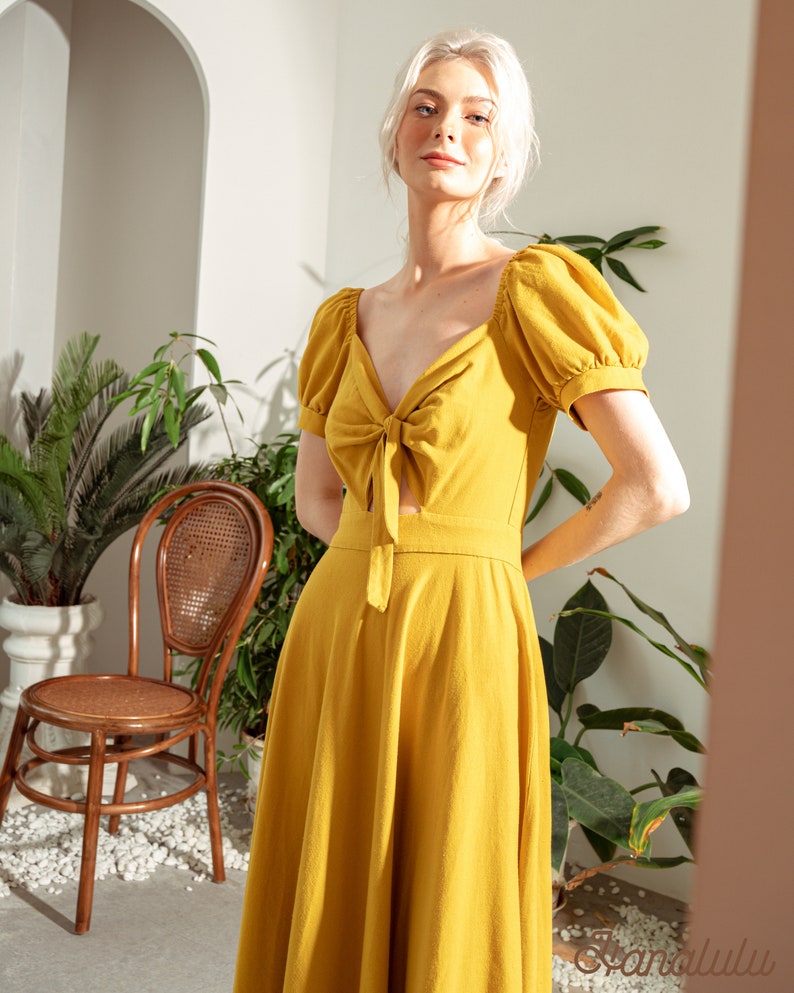 May include: A woman wearing a yellow midi dress with a tie at the neckline and puff sleeves. The dress is made of a lightweight fabric and has a flowy silhouette.