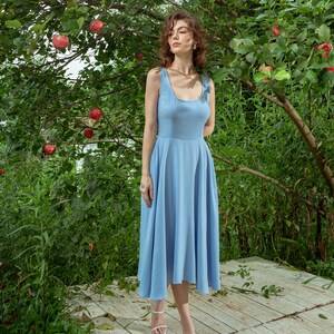 Pu&ograve; includere: Una donna indossa un abito midi senza maniche di colore azzurro chiaro con scollo rotondo e una collana a catena dorata. &Egrave; in piedi su un ponte di legno di fronte a un albero verde con mele rosse.