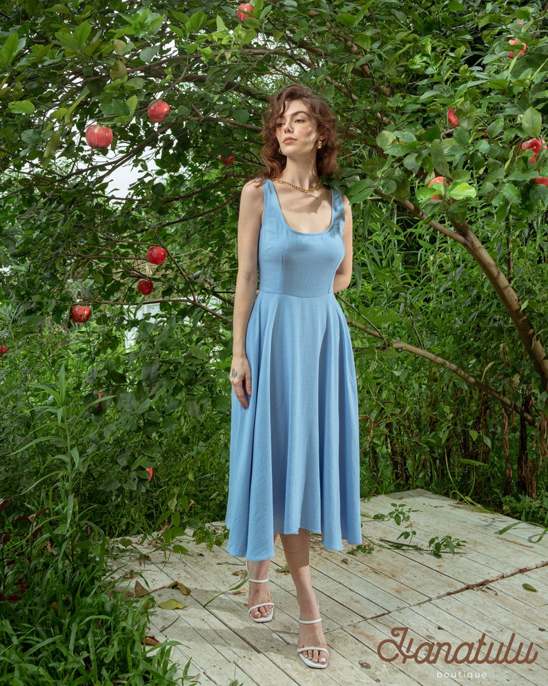 Pu&ograve; includere: Una donna indossa un abito midi senza maniche di colore azzurro chiaro con scollo rotondo e una collana a catena dorata. &Egrave; in piedi su un ponte di legno di fronte a un albero verde con mele rosse.