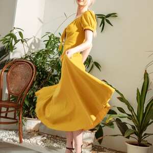 May include: A woman wearing a yellow midi dress with a tie at the neckline and puff sleeves. She is standing in a room with plants and a wicker chair. She is wearing black sandals with laces.