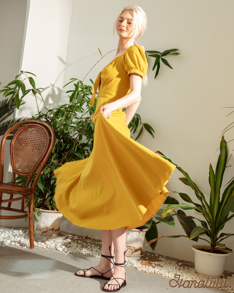May include: A woman wearing a yellow midi dress with a tie at the neckline and puff sleeves. She is standing in a room with plants and a wicker chair. She is wearing black sandals with laces.