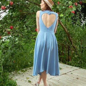 Pu&ograve; includere: Una donna indossa un abito azzurro senza maniche con uno scollo a forma di cuore sulla schiena. &Egrave; in piedi su un ponte di legno con un albero verde e mele rosse sullo sfondo. Indossa un cappello di paglia e tacchi bianchi.