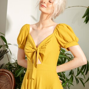 May include: A yellow, short-sleeved dress with a tie at the neckline and a cut-out detail. The dress has a fitted bodice and a flowing skirt.
