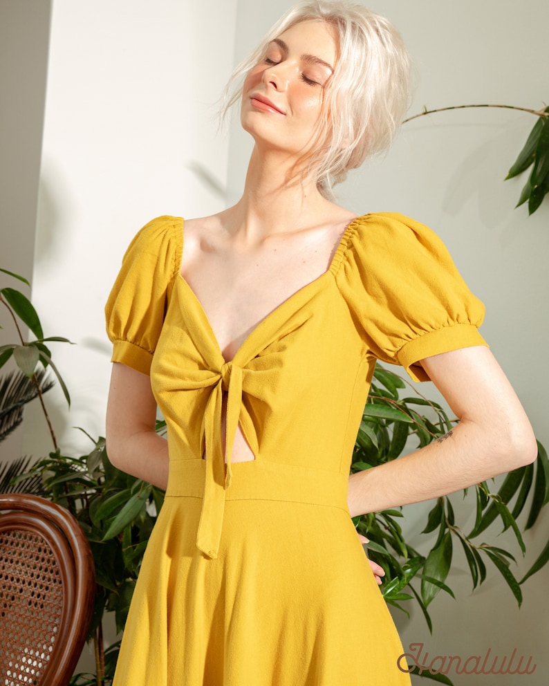 May include: A yellow, short-sleeved dress with a tie at the neckline and a cut-out detail. The dress has a fitted bodice and a flowing skirt.