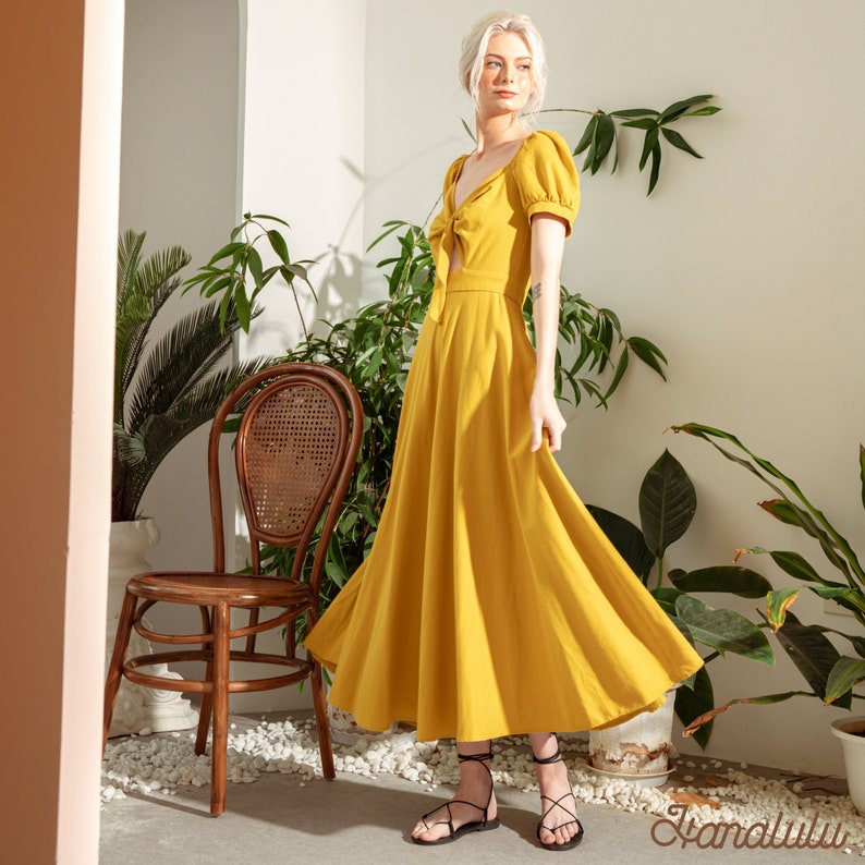 May include: A woman wearing a yellow midi dress with a tie detail at the neckline. The dress has short puff sleeves and a flowy skirt. She is standing in front of a potted plant and a wooden chair.