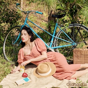 Peut inclure: Une femme en robe pêche est assise sur une couverture dans une zone herbeuse avec un vélo bleu derrière elle. Elle tient un verre de jus et une pomme. Un chapeau de paille et un livre sont sur la couverture.