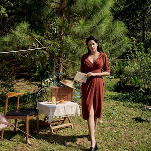 Peut inclure: Une femme en robe rouille se tient dans un espace herbeux avec un livre à la main. Elle porte des chaussures noires et un collier. Il y a une table de pique-nique avec un panier en osier et un vélo en arrière-plan.