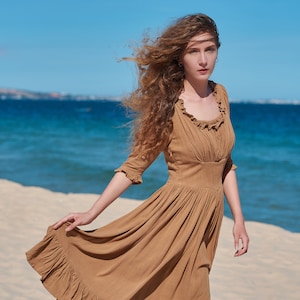 Könnte beinhalten: Eine Frau mit langen, lockigen, braunen Haaren trägt ein braunes, fließendes Kleid mit Rüschen am Ausschnitt. Sie steht an einem Sandstrand mit dem Meer im Hintergrund.