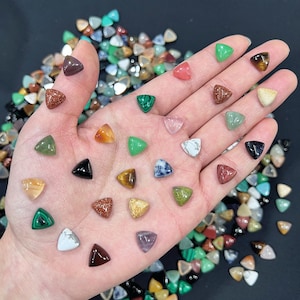 Puede incluir: Una mano sosteniendo una variedad de pequeños cabochones de piedras preciosas triangulares. Los cabochones son de varios colores, incluyendo verde, rojo, azul y marrón. Los cabochones están dispuestos en un patrón en la mano.