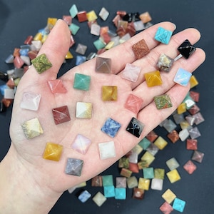 Peut inclure: Une collection de petites pyramides en pierres précieuses colorées. Les pyramides sont faites de différents types de pierres, notamment l'améthyste, le quartz rose et la turquoise. Elles sont disposées dans une main, les pointes des pyramides pointant vers le haut.