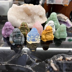 May include: A collection of eight small, carved stone figurines of the Buddha, each in a different color and material. The figurines are arranged in a semi-circle on a black surface.