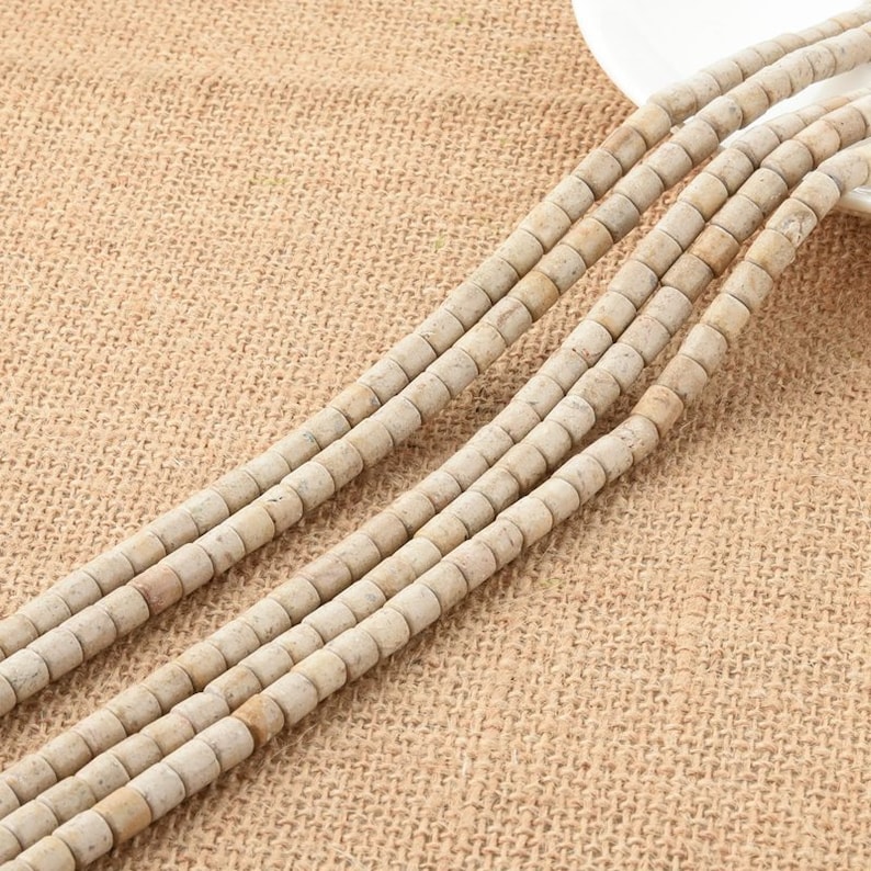 May include: A strand of off-white cylindrical beads made of natural stone. The beads are arranged in rows on a beige burlap background.