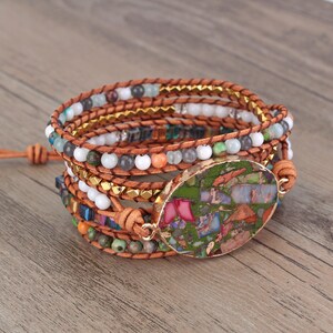 May include: Multi-strand leather wrap bracelet featuring a large oval gemstone pendant. The gemstone is green, brown, and red with a speckled pattern. The bracelet is crafted with brown leather cord, gold beads, and assorted coloured beads.