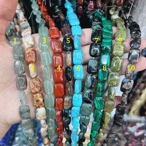 Może przedstawiać: Dziesięć sznurów koralików z kamieni szlachetnych, każdy sznur z innym kolorem i wzorem. Korale są prostokątne i mają gładkie, polerowane wykończenie. Kolory obejmują zielony, niebieski, czerwony, brązowy i czarny.