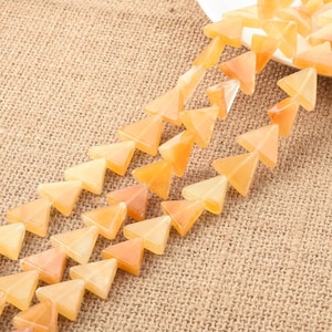 May include: Rows of orange-yellow triangular beads are arranged diagonally on a textured, tan surface. The beads have a translucent quality, with variations in color from pale peach to deeper orange. Some beads are piled on a white plate.