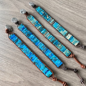 May include: Four wrap bracelets featuring rectangular blue and turquoise beads. The bracelets are connected by brown leather and secured with a silver button clasp. The bracelets are displayed on a wooden surface.