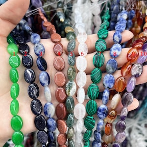 Puede incluir: Diez hebras de cuentas ovaladas de piedras preciosas en varios colores, incluyendo verde, azul, marrón, blanco y rojo. Las cuentas están dispuestas en una fila en una mano.