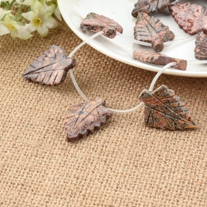 May include: A strand of brown and red leaf-shaped gemstone beads on a clear string. The beads are carved from a stone with a mottled pattern.