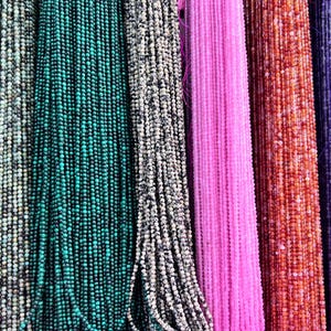 May include: Assortment of colourful gemstone beads strung together in long strands. The beads are various shades of grey, teal, white, pink, orange, and purple.