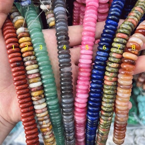 Puede incluir: Una mano sostiene ocho hilos de cuentas de piedras preciosas de varios colores. Las cuentas son planas, redondas y están ensartadas. Los colores incluyen rojo, amarillo, verde, gris, rosa, azul, verde-marrón y naranja. Las cuentas tienen aproximadamente 0,6 cm de diámetro.