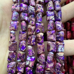 Puede incluir: Una hebra de cuentas de piedras preciosas con patrones morados y blancos. Las cuentas son de forma cuadrada y tienen un acabado pulido.