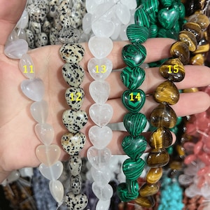 May include: A strand of heart-shaped gemstone beads in various colors and patterns. The beads are strung on a clear cord and are numbered 11 through 15. The beads are made of white, black, green, and brown gemstones.