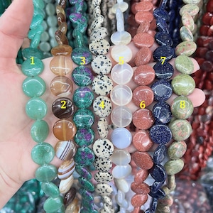 May include: A selection of gemstone beads in various colours and patterns. The beads are round and flat, strung together to form strands. Colours include green, brown, black and white, red, and blue. The beads are held in a hand.