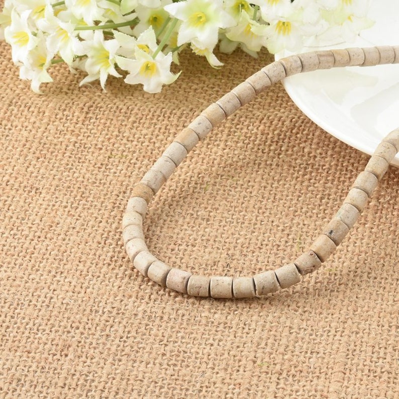 May include: A strand of off-white round beads made of natural stone. The beads are strung on a clear thread and are arranged in a circular pattern. The beads are lying on a brown burlap surface.
