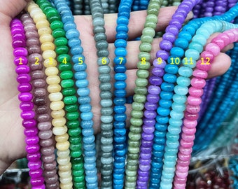 May include: Twelve strands of round, smooth beads in various shades of blue, green, purple, and pink. The beads are strung on a clear thread and are displayed on a hand.