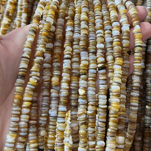 Może przedstawiać: Zbliżenie na wiele sznurów małych, płaskich, dyskowatych koralików. Koraliki są w odcieniach żółci, brązu i bieli. Są nawleczone razem, tworząc teksturowany wygląd. Koraliki są prawdopodobnie wykonane z kamienia naturalnego lub muszli.