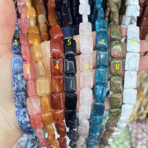 Może przedstawiać: Dziewięć sznurów kwadratowych koralików z kamieni szlachetnych. Koraliki są w różnych kolorach, w tym niebieskim, czerwonym, pomarańczowym, różowym, czarnym, białym i zielonym. Koraliki są nawleczone na sznurek i gotowe do użycia do tworzenia biżuterii.