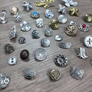 May include: Assortment of silver and gold metal buttons with various designs, including flowers, hearts, trees, and geometric shapes. Some buttons have a textured finish.