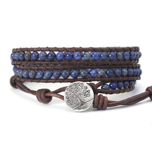 Könnte beinhalten: Ein braunes Leder-Wickelarmband mit blauen Lapislazuli-Perlen und einem silbernen Lebensbaum-Anhänger.